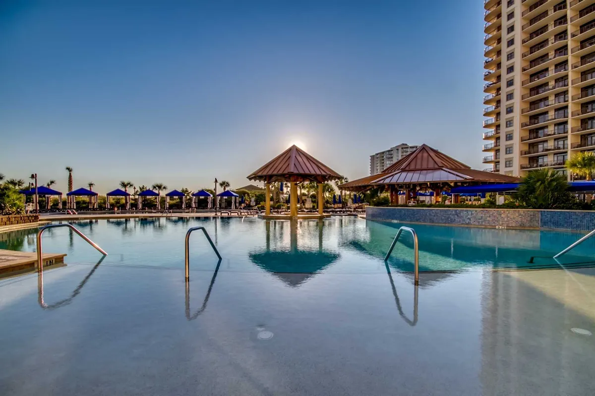 A luxurious resort pool scene with thatched-roof cabanas, inviting lounge chairs, and sunset lighting. The atmosphere feels tranquil and exclusive, perfect for relaxation or socializing near the water.