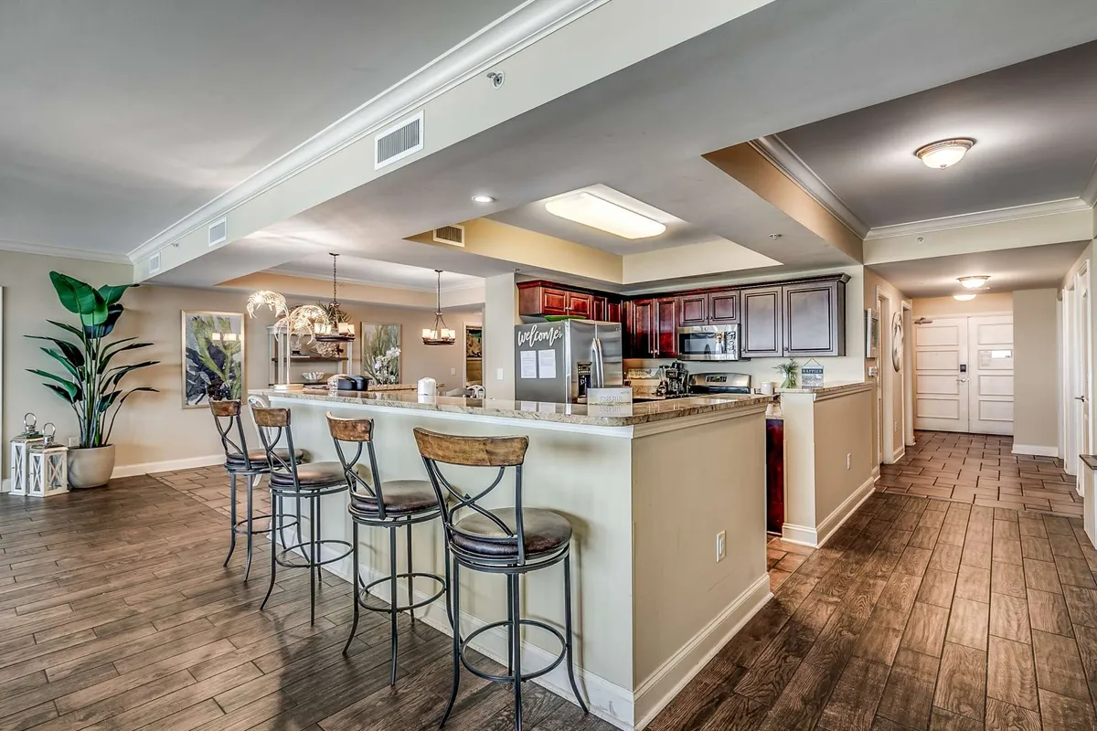 The photo features a kitchen island with bar stools, dark wooden cabinets, and large windows flooding the space with natural light. The setup encourages casual dining, morning coffee rituals, or gathering around the island while meals are prepared.