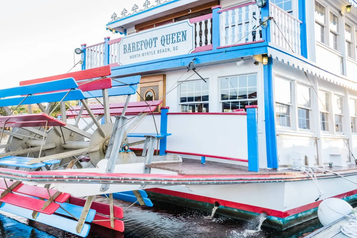 The colorful exterior of the Barefoot Queen riverboat, with its classic paddlewheel, lively signage, and a festive atmosphere. The boat is docked and ready for sightseeing cruises and family gatherings.