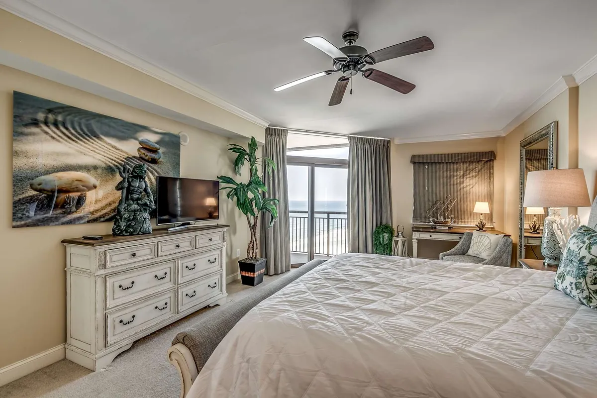 A guest bedroom with a large bed draped in a quilted cover, complemented by stylish artwork on the wall. The room features a television for entertainment, a dressing table, and a large window that fills the space with natural light and offers an inviting view.