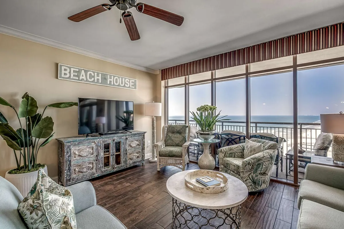 This image features a casual and inviting media room, complete with a TV, comfortable furniture arrangement, decorative plant, and coffee table. Large windows offer a beach view, flooding the room with natural light and connecting the indoor relaxation space with the outdoors.