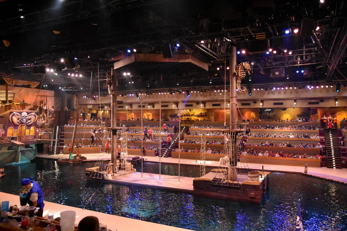 A lively, pirate-themed stage show in progress, with dramatic set designs including a water tank, interactive props, and an audience captivated by the performance. The mood is festive and theatrical, suggesting a memorable entertainment experience.