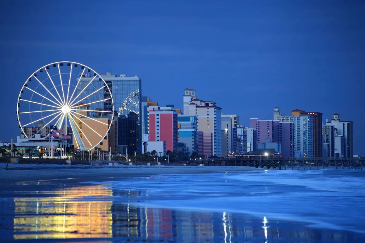 An exterior night view featuring a vibrant Ferris wheel, colorful waterfront buildings, and the ocean’s reflection. The whimsical, celebratory atmosphere suggests family fun, amusements, and shared adventures.