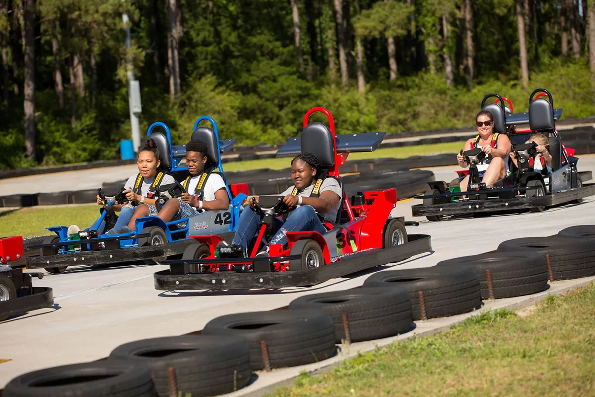 An outdoor go-kart track filled with participants racing brightly colored karts. The scene is energetic and action-packed, with families and friends enjoying the thrill of friendly competition, set amid a backdrop of lush trees.