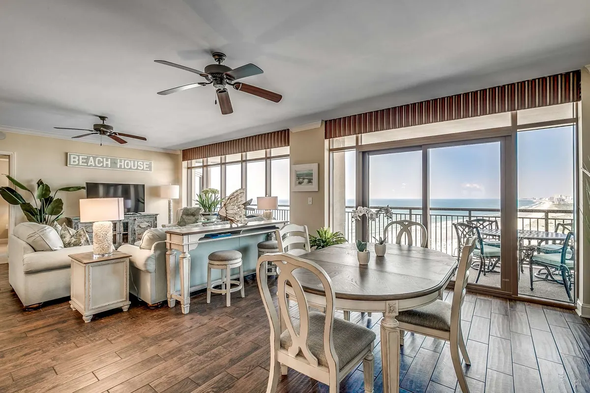 This interior image showcases a spacious, open-plan living and dining area filled with natural light from large windows. The dining table and comfortable seating area are oriented to take in sweeping ocean views, and the decor includes a beach house sign and tasteful accents that create a welcoming, luxurious atmosphere.
