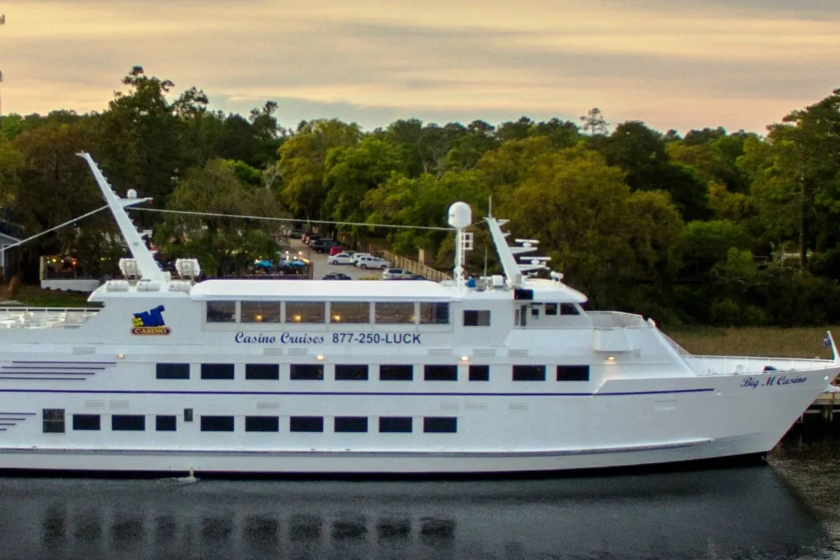 A lively exterior shot of the large white 'Big M Casino' yacht docked amidst greenery and water. The scene evokes excitement and the promise of entertainment, dining, and group outings.