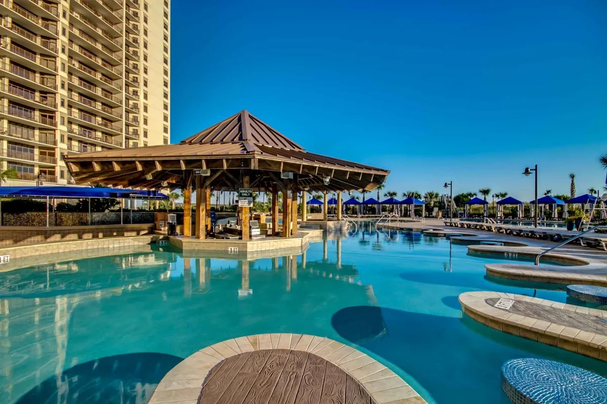 A vibrant outdoor pool area featuring a large swimming pool surrounded by palm trees, sun loungers, blue umbrellas, and a poolside cabana. The setting is lively with guests socializing, swimming, and soaking up the sun, capturing the resort's expansive pool paradise.