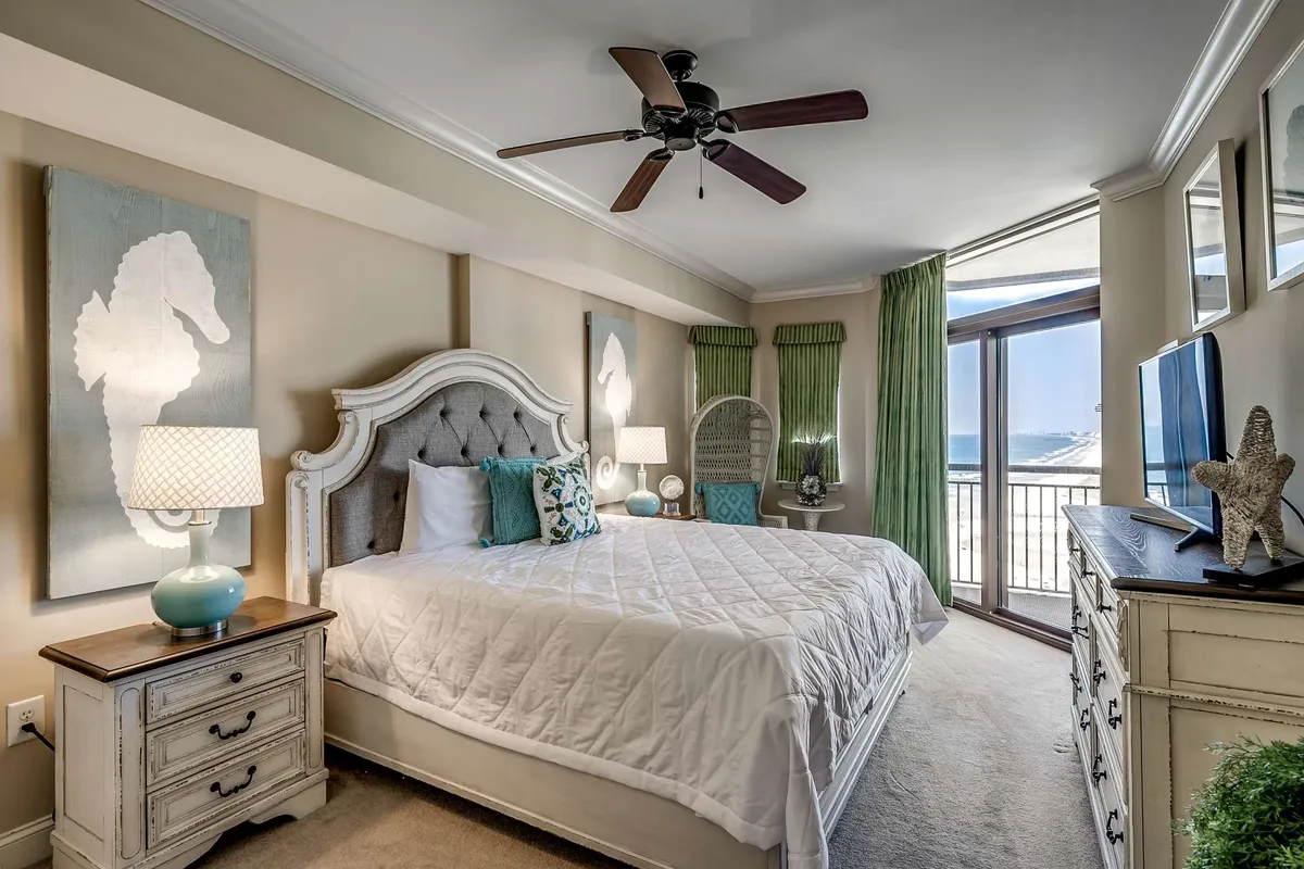 A spacious master bedroom featuring a king-sized bed with crisp white bedding, accented by teal and blue decorative pillows. The space is bathed in natural light from a large window that offers a sweeping beach view. Two seahorse wall art pieces and a dresser with decorative items complete the serene, coastal-inspired decor.